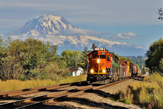 The FRA continues to focus on Positive Train Control (PTC) implementation with a new push to speed progress as well as the release of more grant funding.  A BNSF train speeds along main line track that will be part of the PTC network near Puyallup, Washington beneath Mount Rainier.  Image © 8/2016 by Nikki Burgess; all rights reserved.